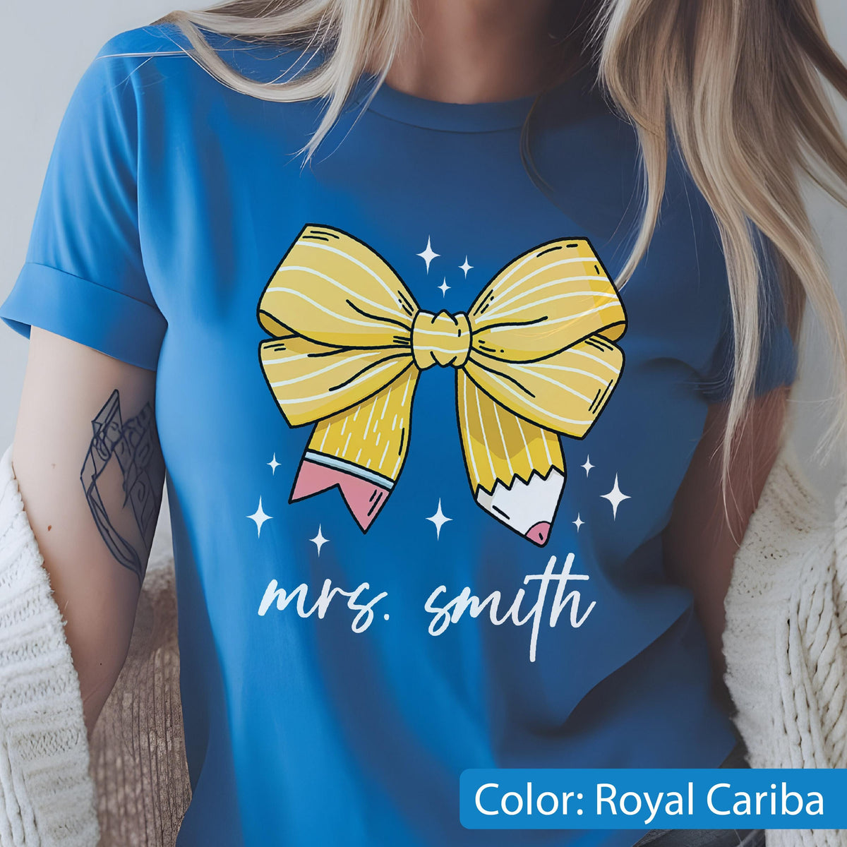 May include: Royal Cariba blue t-shirt featuring a yellow bow design with pencil details and the text "mrs. smith" in white script. The bow has yellow stripes and is surrounded by white star accents. The shirt is a solid color.