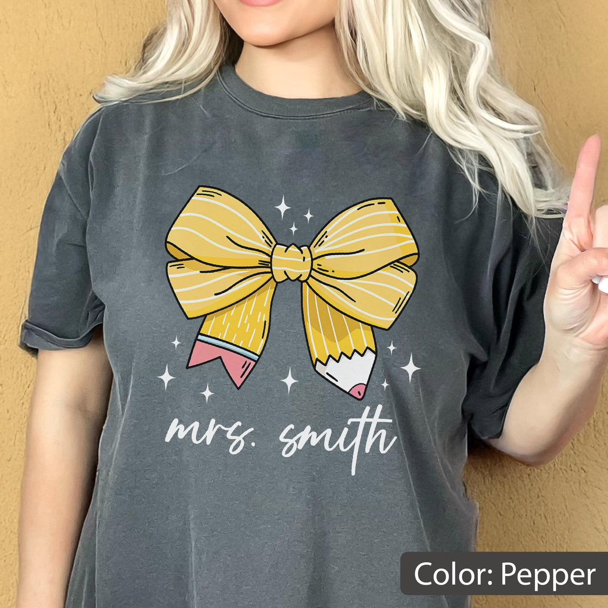 May include: Pepper-colored t-shirt featuring a large yellow bow designed as a pencil. The bow is centered on the shirt, with the text "mrs. smith" in a cursive font below it. Small white stars are scattered around the bow.