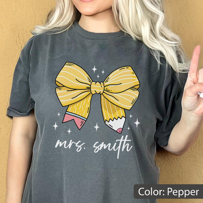 May include: Pepper-colored t-shirt featuring a large yellow bow designed as a pencil. The bow is centered on the shirt, with the text "mrs. smith" in a cursive font below it. Small white stars are scattered around the bow.