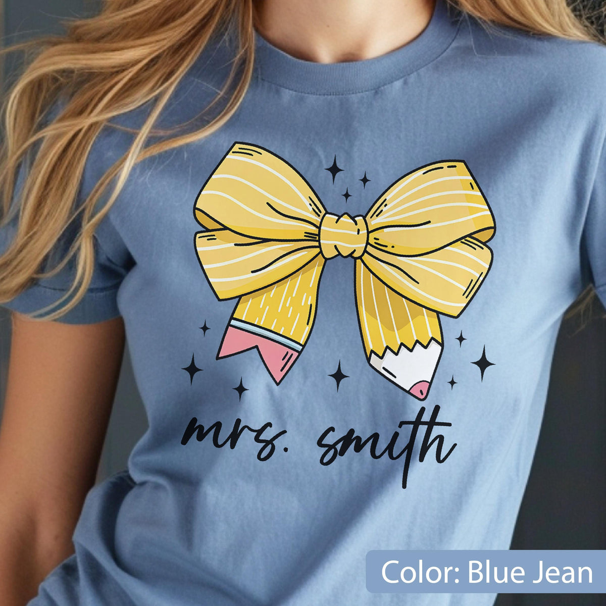 May include: Blue jean-colored t-shirt featuring a large yellow bow design with pencil accents and the text "mrs. smith". The bow has white stripes and is surrounded by small black star shapes. The shirt is made of soft material.