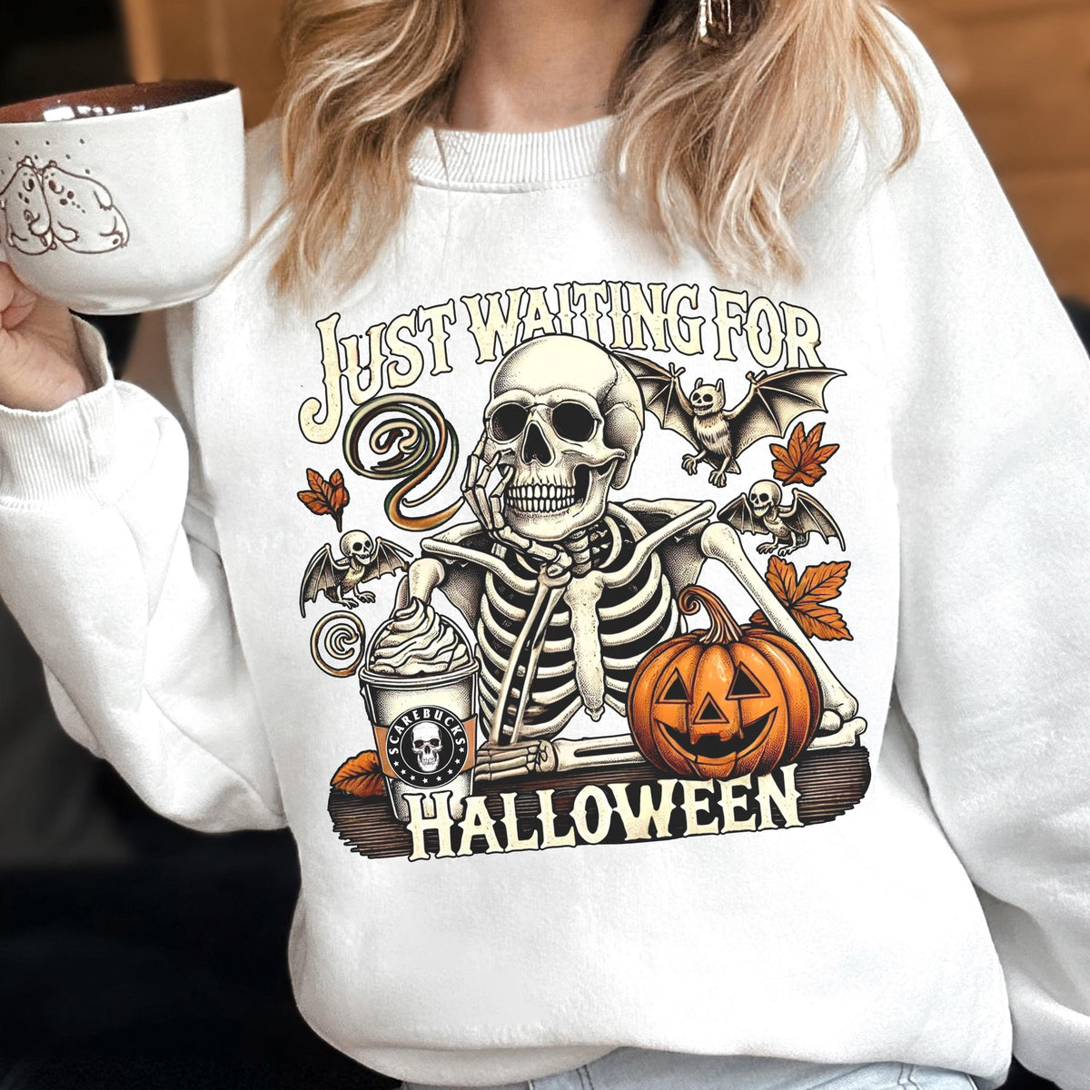 May include: A white crewneck sweatshirt featuring a graphic of a skeleton resting its chin on its hand. The design includes a coffee cup labeled 'Scarebucks', a jack-o'-lantern, bats, autumn leaves, and the text 'JUST WAITING FOR HALLOWEEN'.
