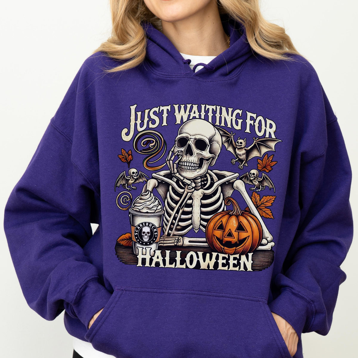 May include: A purple hooded sweatshirt featuring a graphic of a skeleton resting its chin on its hand next to a coffee cup and a pumpkin. The text reads "JUST WAITING FOR HALLOWEEN" with illustrations of bats and autumn leaves surrounding the skeleton.