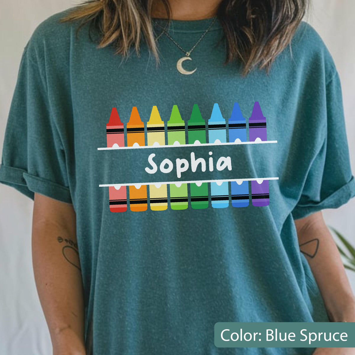 May include: Blue spruce t-shirt with a colorful crayon design. The design includes a row of crayons above and below the name "Sophia" in white. The crayons are in rainbow colors.