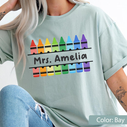 May include: A light green t-shirt featuring a colorful crayon design with the text "Mrs. Amelia" in black. The crayons are arranged in a rainbow pattern. The shirt is a solid color and has rolled-up sleeves.