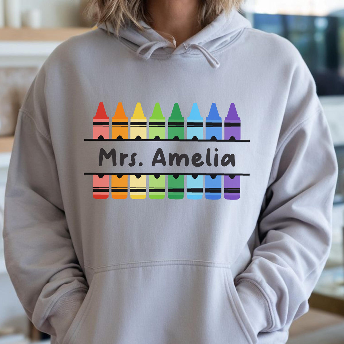 May include: A light gray hoodie with a colorful crayon design. The design includes a row of crayons in rainbow colors above and below the text "Mrs. Amelia". The hoodie has a front pocket and a hood.