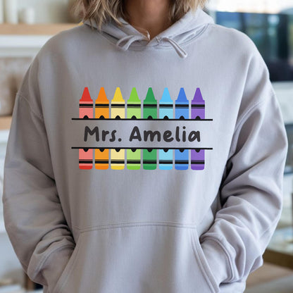 May include: A light gray hoodie with a colorful crayon design. The design includes a row of crayons in rainbow colors above and below the text "Mrs. Amelia". The hoodie has a front pocket and a hood.