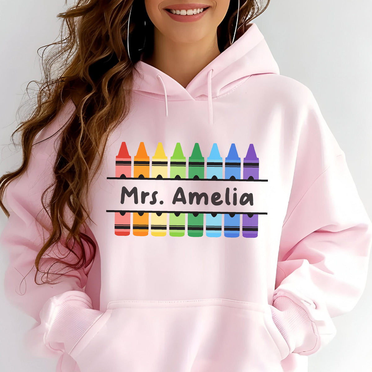 May include: Light pink hoodie with a crayon design. The design features a row of colorful crayons above and below the text "Mrs. Amelia". The crayons are in a rainbow color scheme. The hoodie has a drawstring and a front pocket.