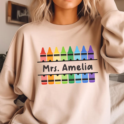 May include: Beige sweatshirt featuring a colorful crayon design with the text "Mrs. Amelia" in black. The crayons are arranged in a rainbow pattern. The sweatshirt is a neutral color.