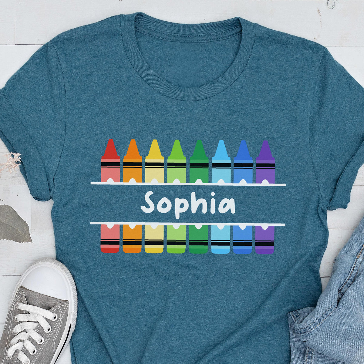 May include: Teal t-shirt featuring a colorful crayon design with the name "Sophia" in white. The design includes a row of crayons above and below the name, each in a different color.