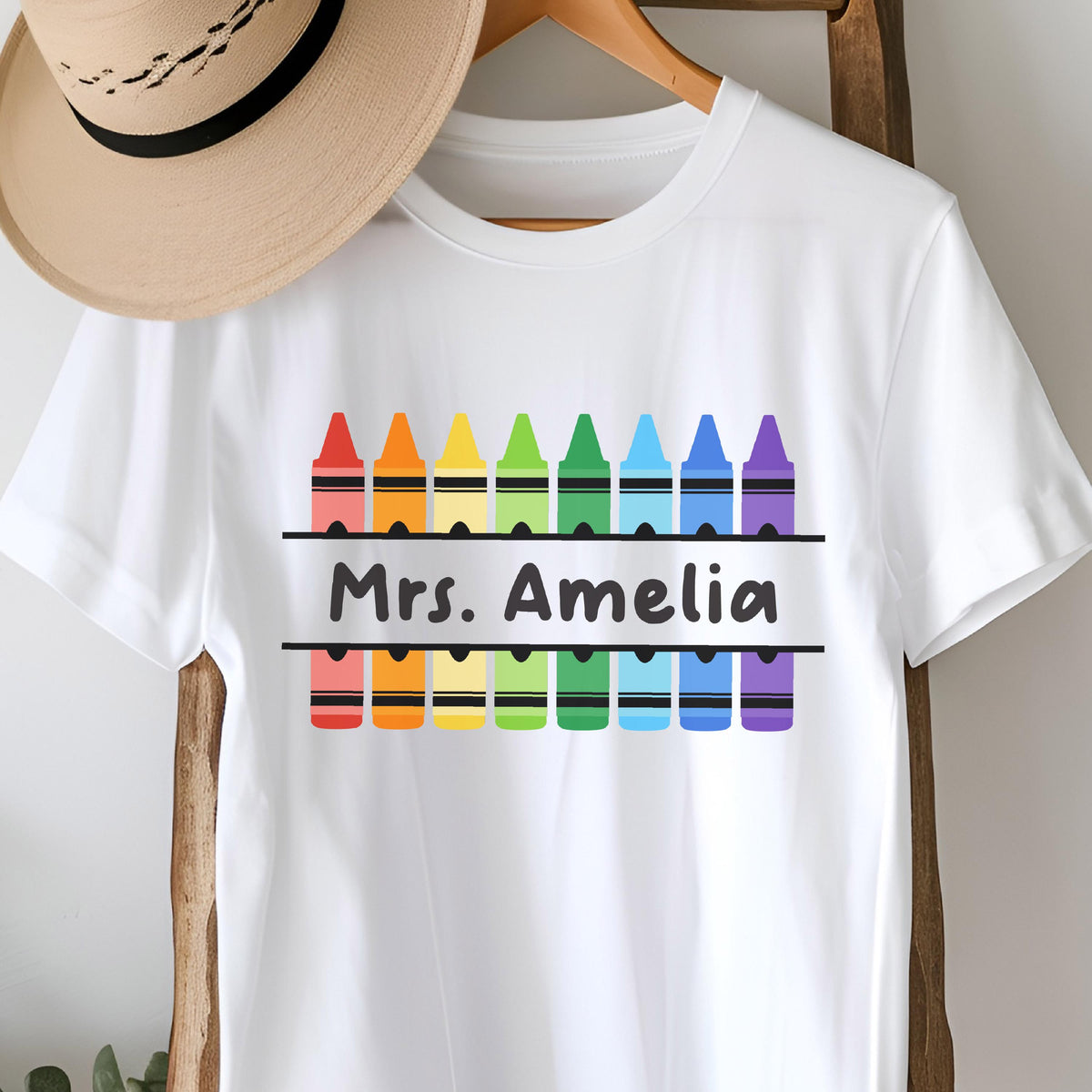 May include: White t-shirt featuring a colorful crayon design above and below the text "Mrs. Amelia". The crayons are arranged in a rainbow pattern. The shirt is displayed on a wooden hanger.