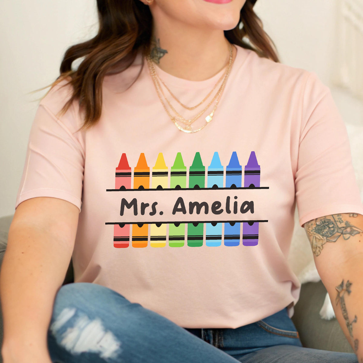 May include: A light pink t-shirt featuring a colorful crayon design. The design includes a row of crayons in rainbow colors above and below the text "Mrs. Amelia" in black. The shirt is a crew neck style.