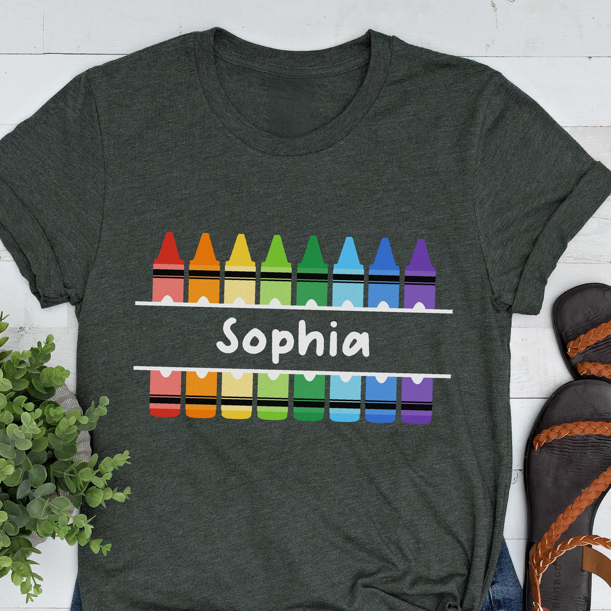 May include: Dark gray t-shirt with a colorful crayon design. The design includes a row of crayons above and below the name "Sophia" in white, with a rainbow of colors. The shirt is a casual style.