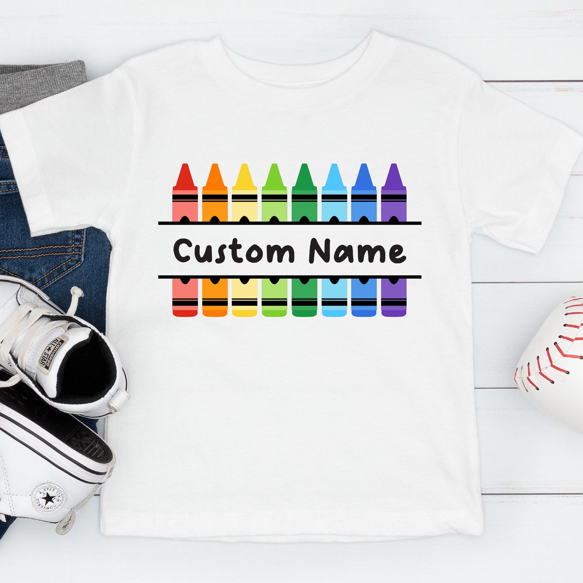 May include: A white short-sleeved t-shirt featuring a graphic of two rows of colorful crayons in red, orange, yellow, green, blue, and purple. Between the rows of crayons, the text Custom Name is printed in a bold black sans-serif font.