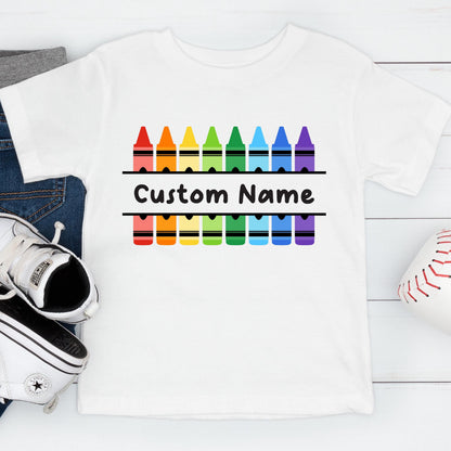 May include: A white short-sleeved t-shirt featuring a graphic of two rows of colorful crayons in red, orange, yellow, green, blue, and purple. Between the rows of crayons, the text Custom Name is printed in a bold black sans-serif font.