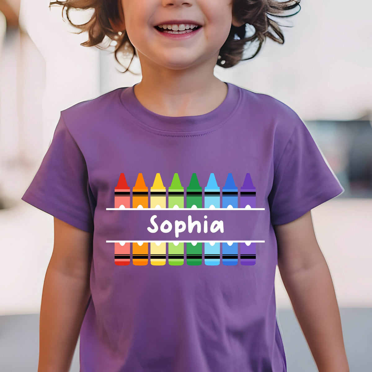 May include: A purple short-sleeved t-shirt featuring a graphic of two rows of colorful crayons in red, orange, yellow, green, light blue, dark blue, and purple. Between the rows is a white horizontal bar with the name Sophia printed in white.