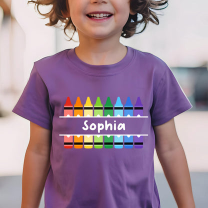 May include: A purple short-sleeved t-shirt featuring a graphic of two rows of colorful crayons in red, orange, yellow, green, light blue, dark blue, and purple. Between the rows is a white horizontal bar with the name Sophia printed in white.