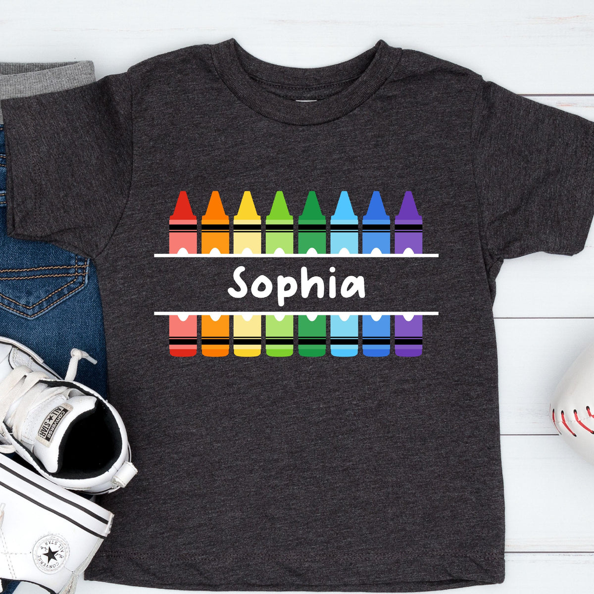May include: A dark charcoal gray short-sleeve t-shirt featuring a graphic of two rows of colorful crayons in red, orange, yellow, green, blue, and purple. Between the rows of crayons, the name Sophia is printed in white, bold, sans-serif text.