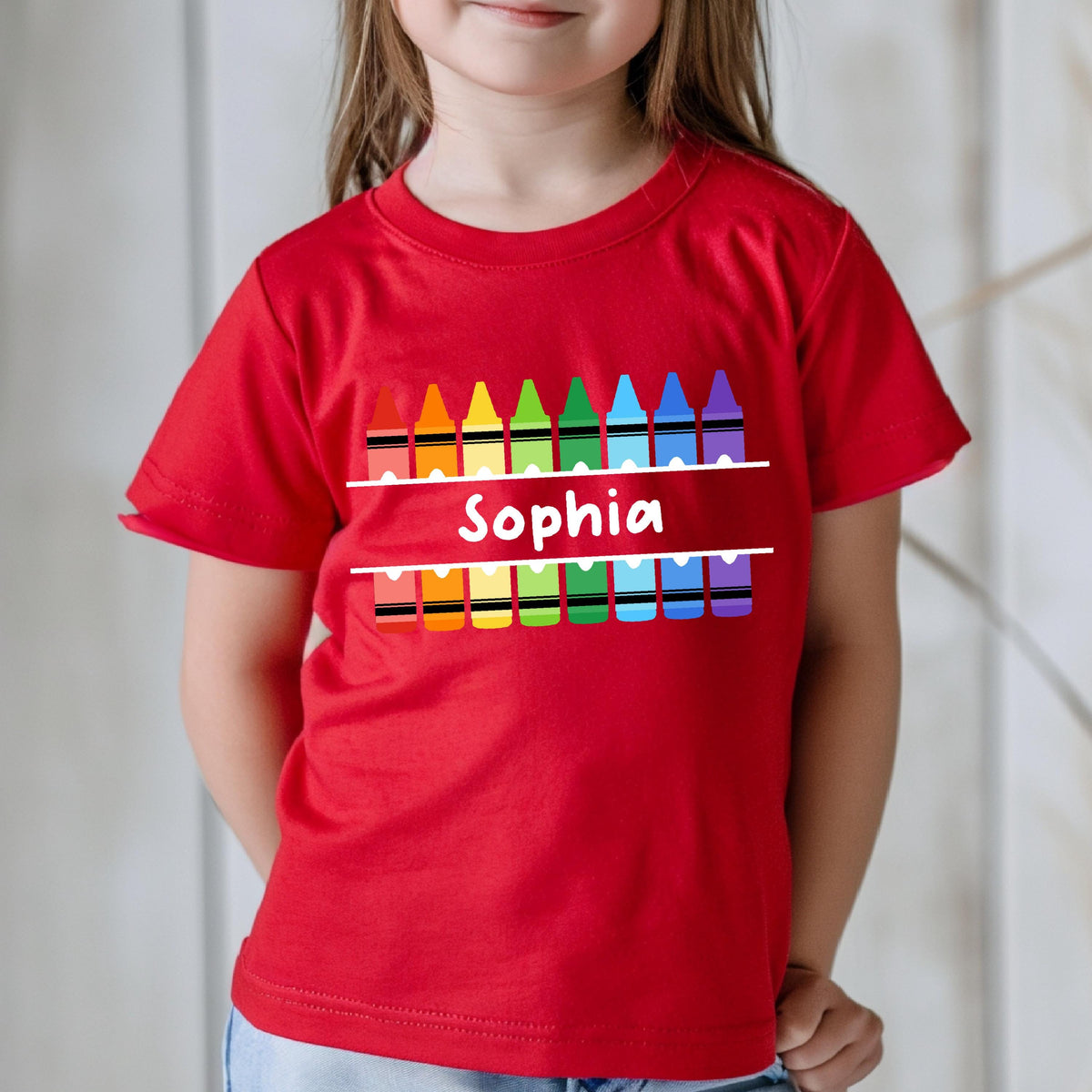 May include: A bright red short-sleeved t-shirt featuring a graphic of two rows of colorful crayons in red, orange, yellow, green, light blue, dark blue, and purple. The name Sophia is printed in white text between the two rows of crayons.