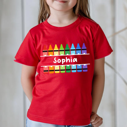May include: A bright red short-sleeved t-shirt featuring a graphic of two rows of colorful crayons in red, orange, yellow, green, light blue, dark blue, and purple. The name Sophia is printed in white text between the two rows of crayons.