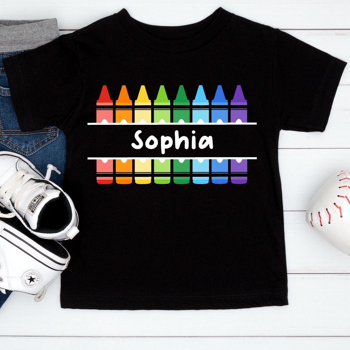 May include: A black short-sleeve t-shirt featuring a graphic of two rows of colorful crayons in rainbow order. Between the rows of crayons, a white horizontal bar contains the name Sophia printed in a bold, white, sans-serif font.