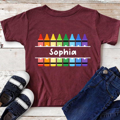 May include: A maroon short-sleeved t-shirt featuring a graphic of two rows of colorful crayons in red, orange, yellow, green, light blue, dark blue, and purple. Between the rows of crayons, the name Sophia is printed in white text.