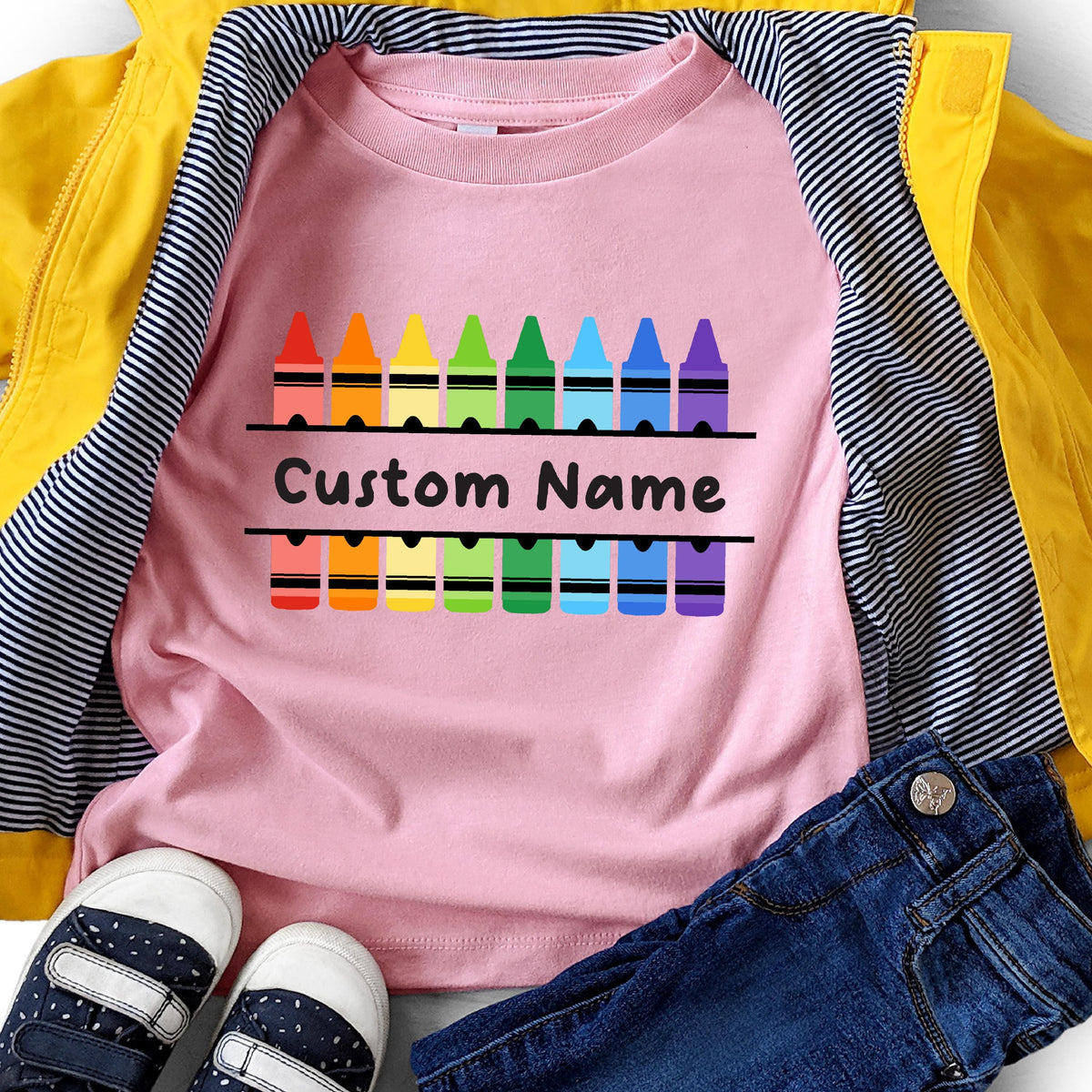 May include: A light pink t-shirt featuring a graphic of two rows of colorful crayons in red, orange, yellow, green, light green, blue, dark blue, and purple. The text Custom Name is printed in black between the two rows of crayon illustrations.