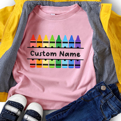 May include: A light pink t-shirt featuring a graphic of two rows of colorful crayons in red, orange, yellow, green, light green, blue, dark blue, and purple. The text Custom Name is printed in black between the two rows of crayon illustrations.