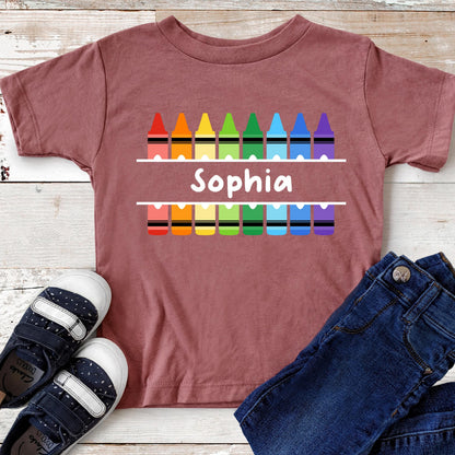 May include: A mauve short-sleeve t-shirt featuring a graphic of two rows of colorful crayons in red, orange, yellow, green, light blue, dark blue, and purple. Between the rows of crayons, the name Sophia is printed in white text.