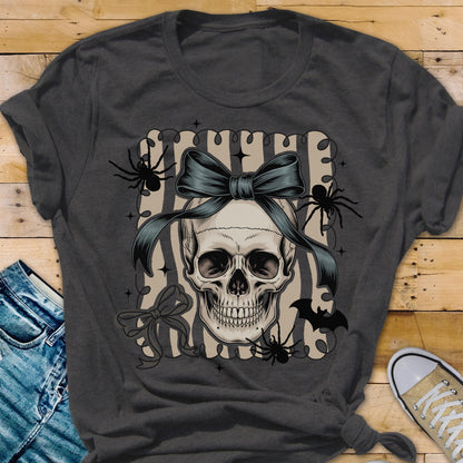 May include: Dark gray t-shirt featuring a skull with a blue bow, spiders, and a bat graphic. The skull is centered on a striped background. A pair of jeans and a sneaker are visible in the image.