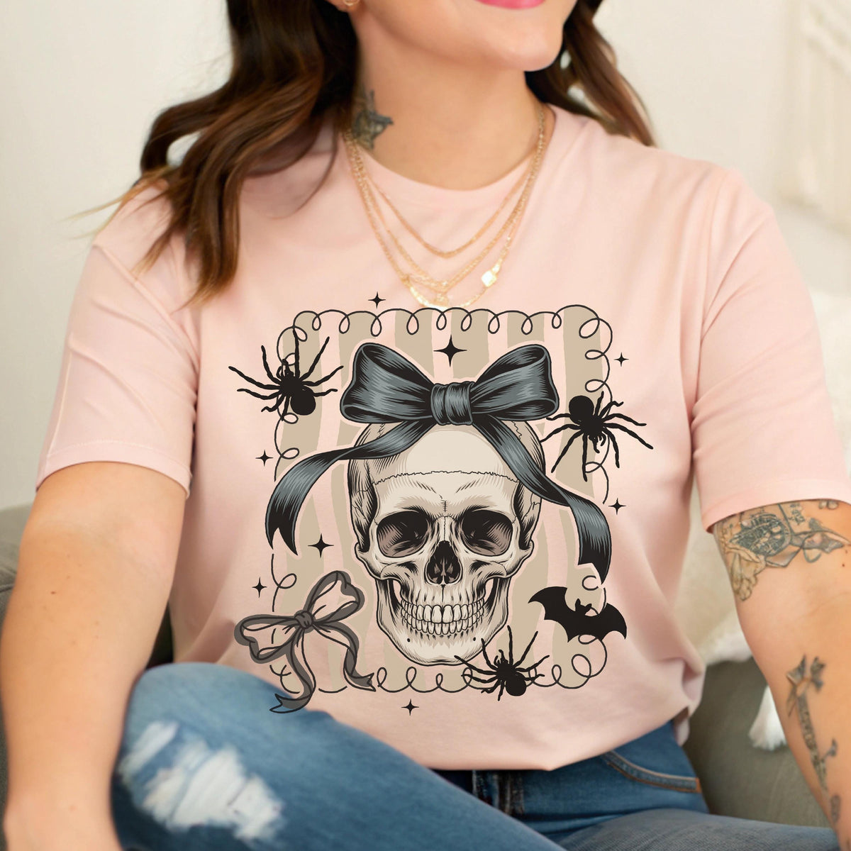May include: A light pink t-shirt featuring a skull with a dark gray bow, surrounded by spiders and a bat. The word "BAD" is above the skull. The design is enclosed in a decorative frame.