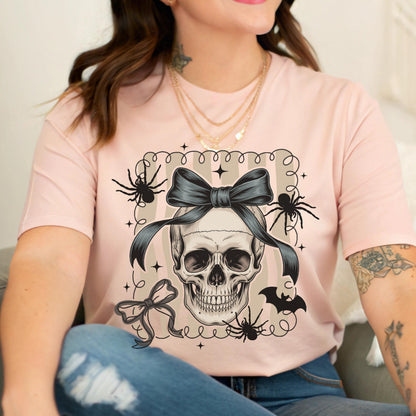 May include: A light pink t-shirt featuring a skull with a dark gray bow, surrounded by spiders and a bat. The word "BAD" is above the skull. The design is enclosed in a decorative frame.
