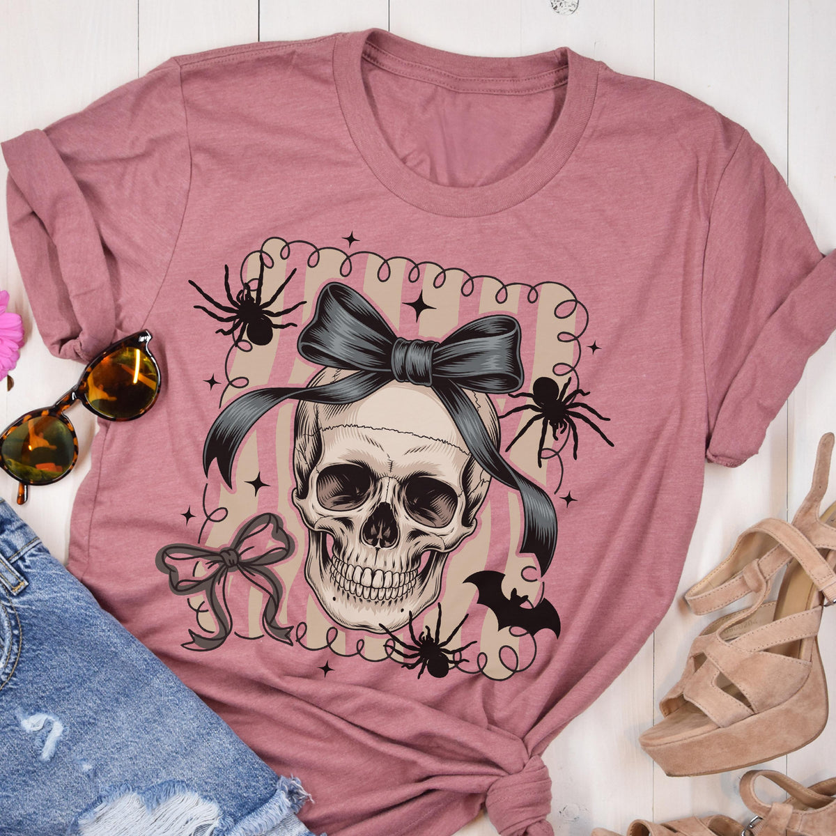 May include: A dusty rose t-shirt featuring a skull with a black bow, spiders, and a bat graphic. The design is framed by a decorative border with stars and a bow. The shirt is a casual wear item.