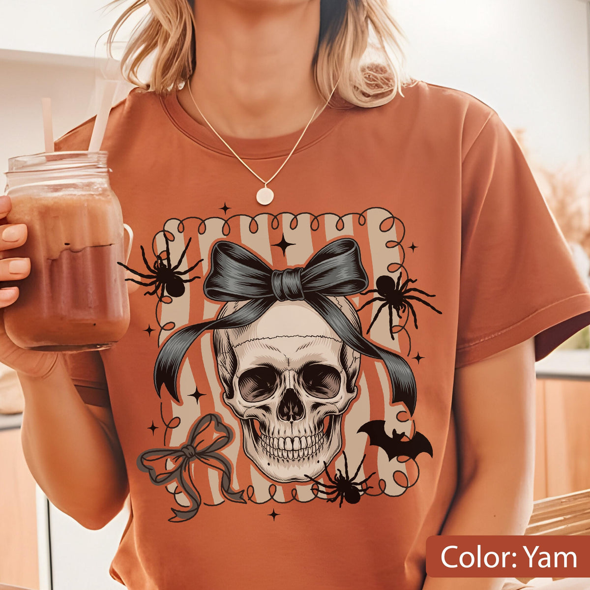 May include: A yam-colored t-shirt featuring a skull design with a black bow, spiders, and a bat. The design is framed with a striped border and stars, creating a Halloween theme.