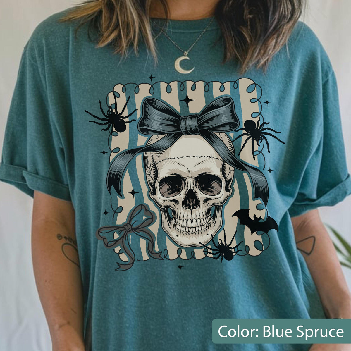 May include: Blue spruce t-shirt featuring a skull design with a bow, spiders, and a bat. The skull is centered within a striped frame. The shirt has rolled-up sleeves.
