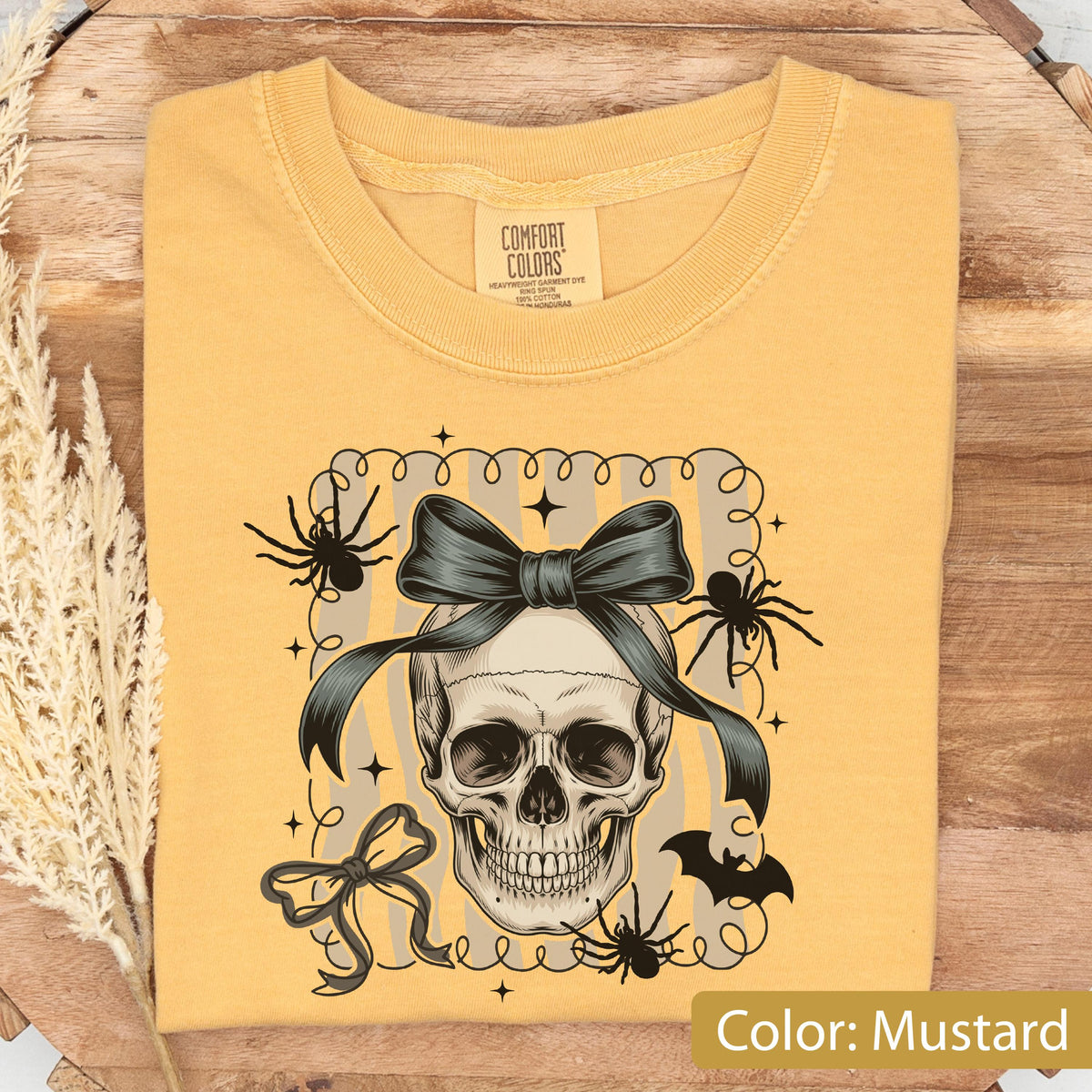 May include: Mustard-colored t-shirt featuring a skull with a dark bow, spiders, and a bat graphic. The shirt has a crew neck and short sleeves. The text "Color: Mustard" is at the bottom right.