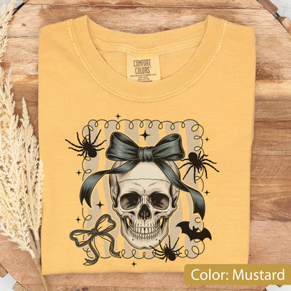 May include: Mustard-colored t-shirt featuring a skull with a dark bow, spiders, and a bat graphic. The shirt has a crew neck and short sleeves. The text "Color: Mustard" is at the bottom right.