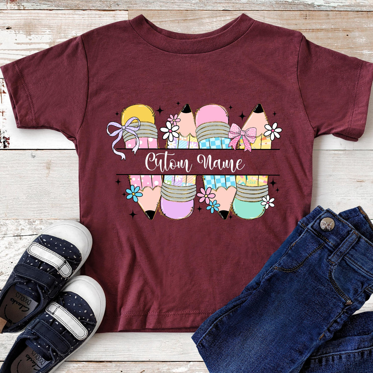 May include: Burgundy t-shirt featuring a colorful pencil design with the text "Custom Name". The pencils are decorated with bows and flowers. A pair of navy blue shoes and blue jeans are visible.
