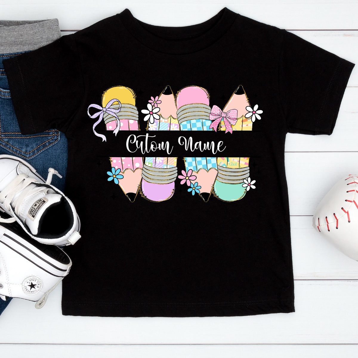 May include: Black t-shirt featuring a colorful pencil design with bows and flowers. The pencils are in shades of yellow, pink, purple, and teal. The text "Custom Name" is displayed across the pencils. A pair of white sneakers and blue jeans are visible.