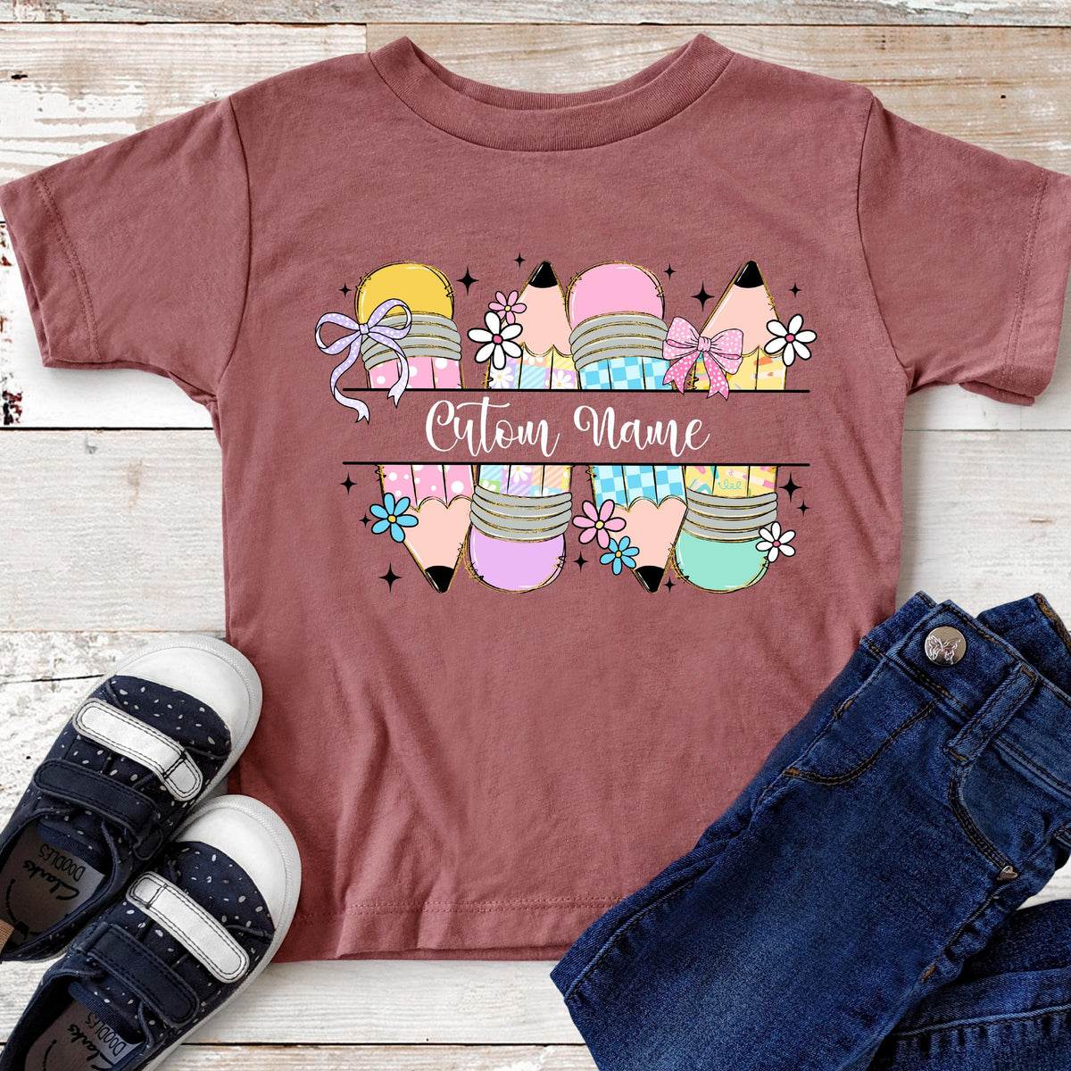 May include: A maroon t-shirt featuring a colorful pencil design with the text "Custom Name". The pencils are decorated with bows and flowers. A pair of navy blue shoes and blue jeans are next to the shirt.