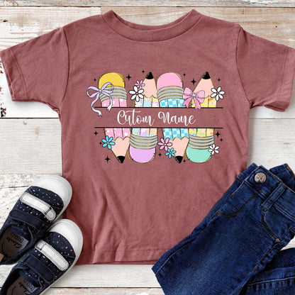 May include: A maroon t-shirt featuring a colorful pencil design with the text "Custom Name". The pencils are decorated with bows and flowers. A pair of navy blue shoes and blue jeans are next to the shirt.