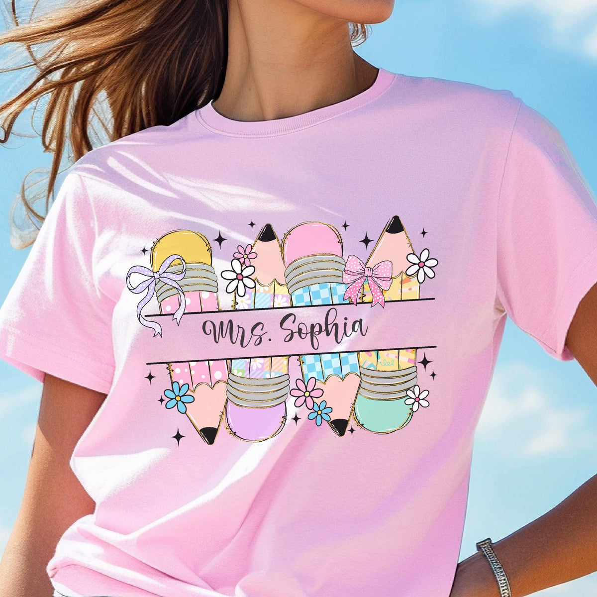 May include: A light pink t-shirt featuring a colorful pencil design with bows and flowers. The text "Mrs. Sophia" is printed in cursive. The pencils are in various colors, including yellow, pink, and blue.