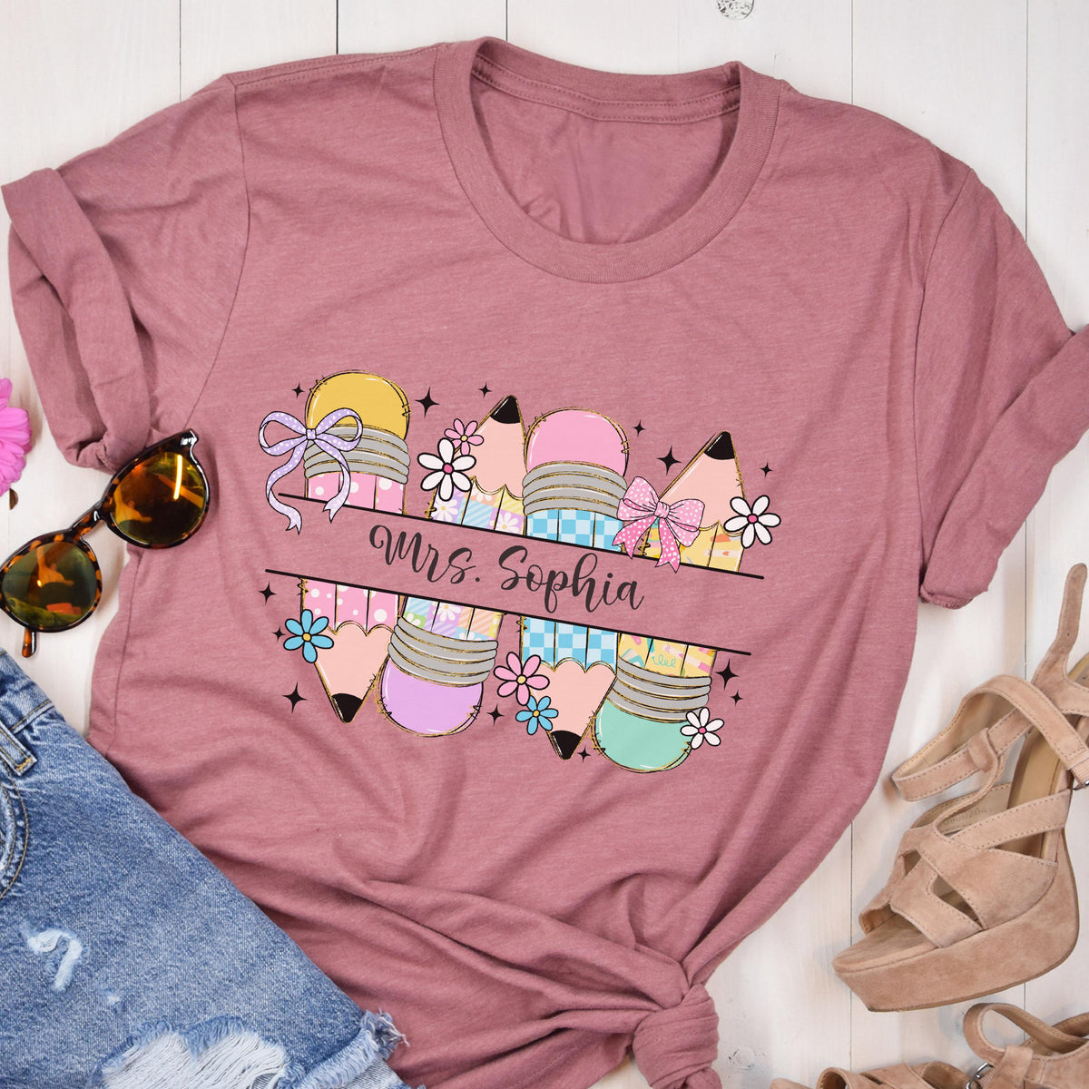May include: Dusty rose t-shirt featuring a colorful pencil graphic with bows and flowers. The text "Mrs. Sophia" is displayed across the pencils. The shirt is a crew neck and has rolled-up sleeves.