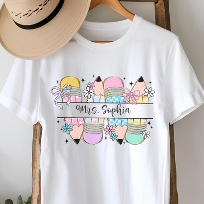 May include: White t-shirt featuring a colorful pencil design with the text "Mrs. Sophia". The pencils are in various colors, including yellow, pink, and teal, with floral and bow accents. A straw hat hangs in the background.