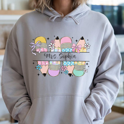 May include: A light gray hoodie featuring a colorful pencil design with bows and flowers. The text "Mrs. Sophia" is printed in a black font. The pencils are in various colors, including yellow, pink, and blue.