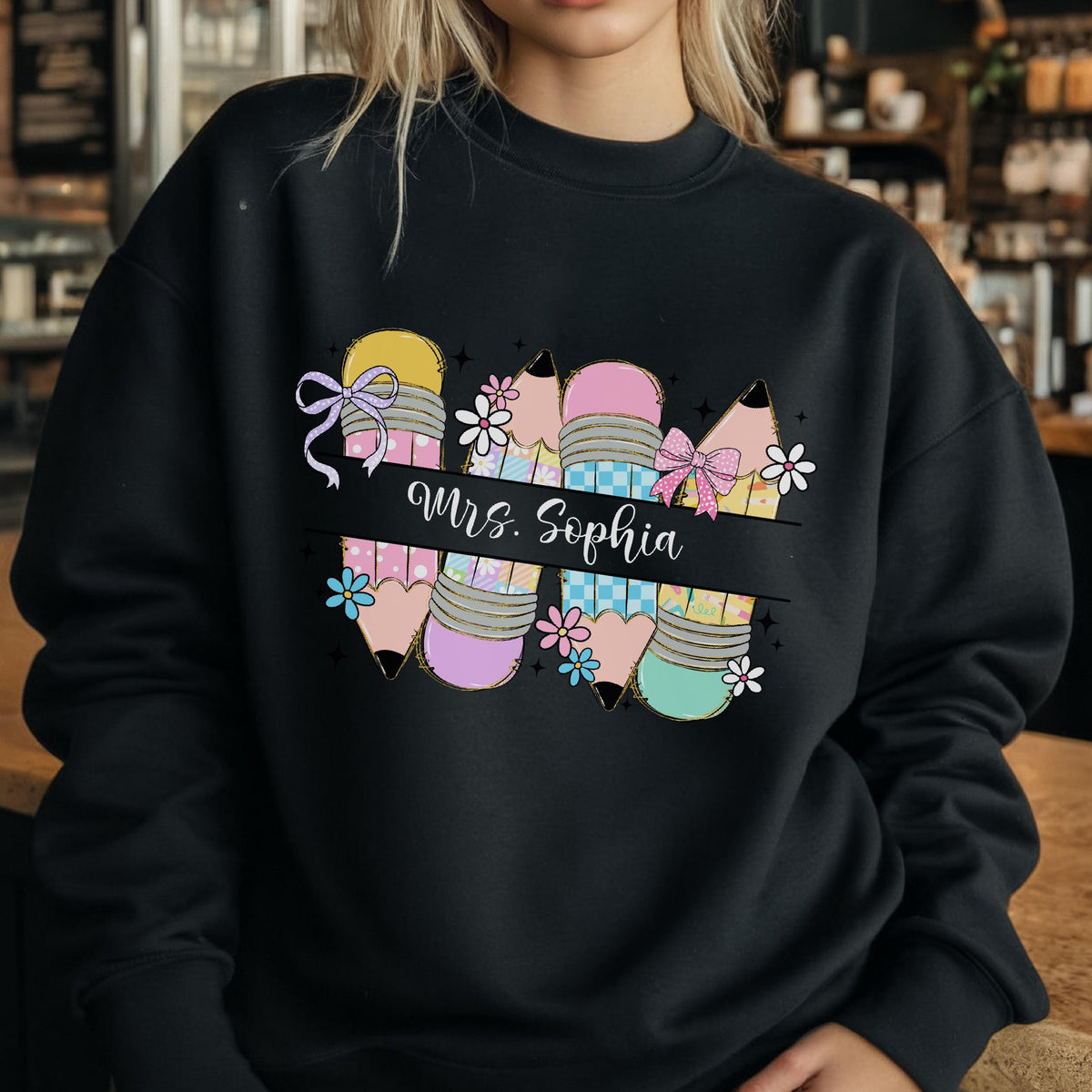 May include: Black sweatshirt featuring a colorful pencil design with the text "Mrs. Sophia". The pencils are decorated with bows, flowers, and patterns in shades of yellow, pink, blue, and green. A perfect gift for teachers.