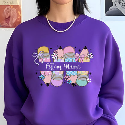 May include: A purple sweatshirt featuring a colorful pencil design with the text "Custom Name". The pencils have various patterns and colors, including pink, yellow, and blue, with floral accents and bows. The sweatshirt is a solid purple color.