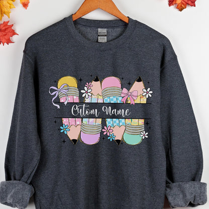 May include: Dark gray sweatshirt featuring a colorful pencil design with bows and flowers. The design includes the text "Custom Name" in a cursive font, perfect for personalization. The pencils are in various pastel colors.