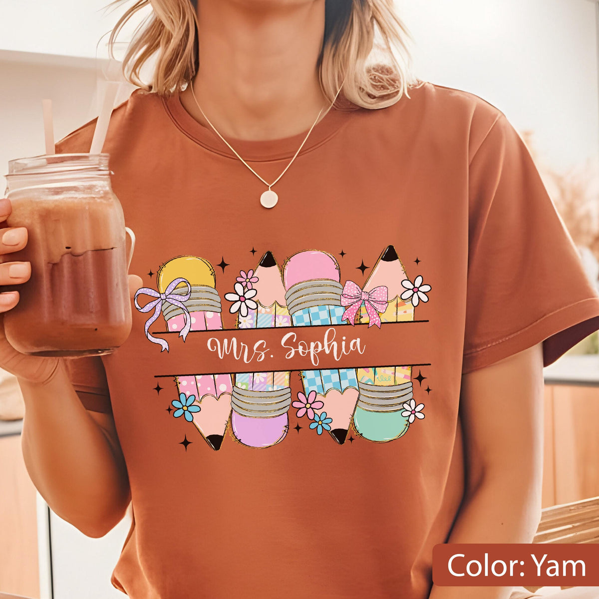 May include: A yam-colored t-shirt featuring a graphic print of four stylized pencils with pastel patterns, small flowers, and bows. The text "Mrs. Sophia" is written in a white script font across a horizontal line centered over the pencil design.