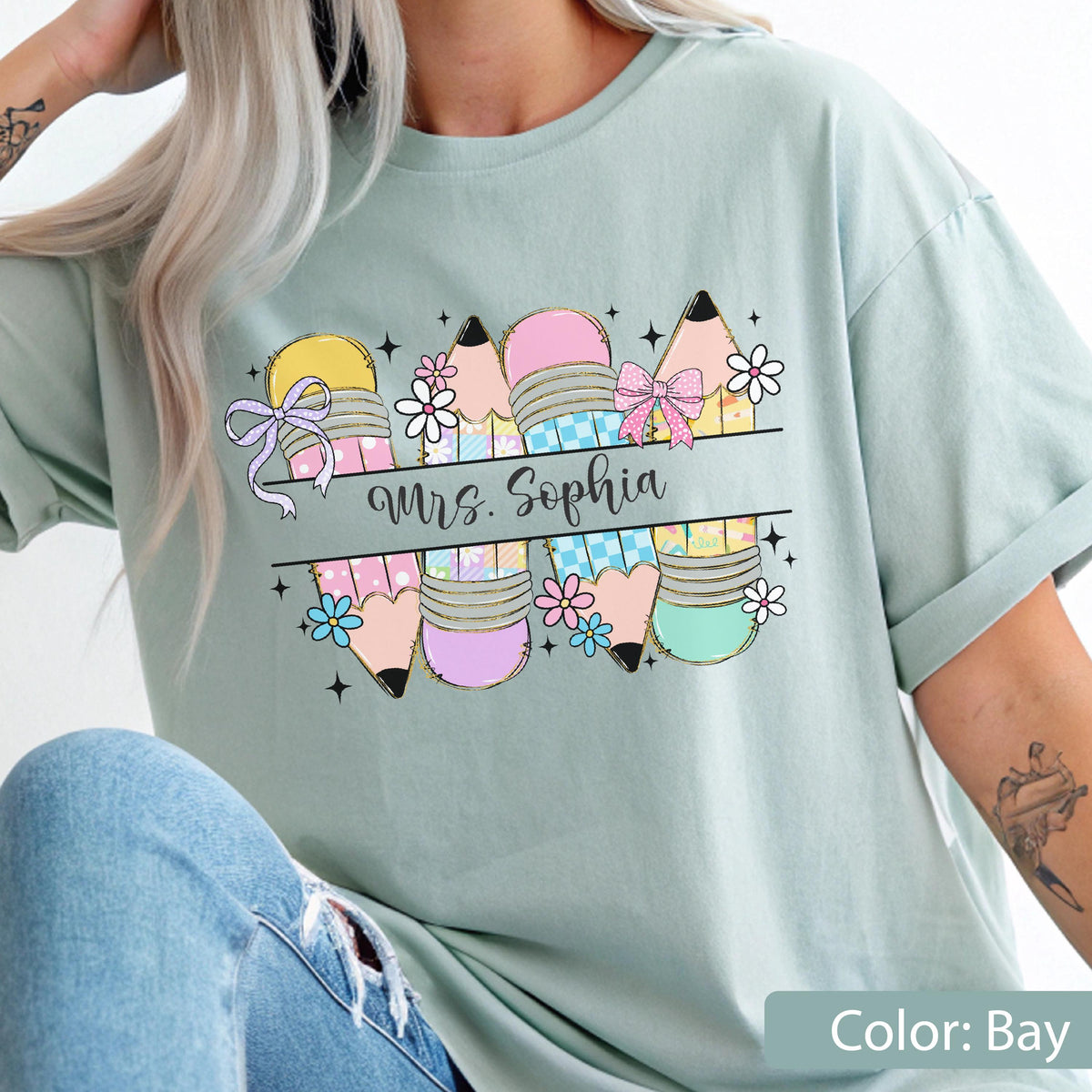 May include: A bay-colored t-shirt featuring a graphic of four pencils with pink, yellow, and blue patterns, decorated with small flowers, bows, and stars. A horizontal banner across the center displays the text Mrs. Sophia in black cursive script.