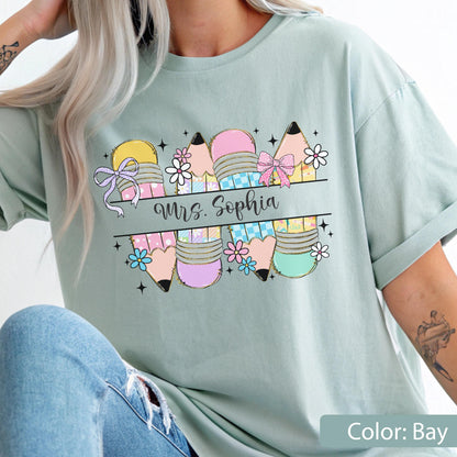 May include: A bay-colored t-shirt featuring a graphic of four pencils with pink, yellow, and blue patterns, decorated with small flowers, bows, and stars. A horizontal banner across the center displays the text Mrs. Sophia in black cursive script.